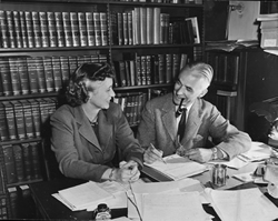 black and white photo or a couple (Annie Burr Lewis and Wilmarth Sheldon Lewis) seated next to each other at a desk, looking at each other, with a wall of books on shelves behind them. 