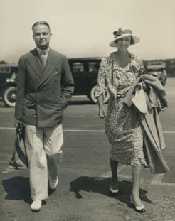 a 1930s black and white photo or W.S. Lewis and Annie Burr Lewis walking side by side toward the camera with a car parked behind them.