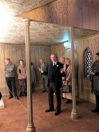 color photo of people listening to man speak in a hallway with gothic wall decoration